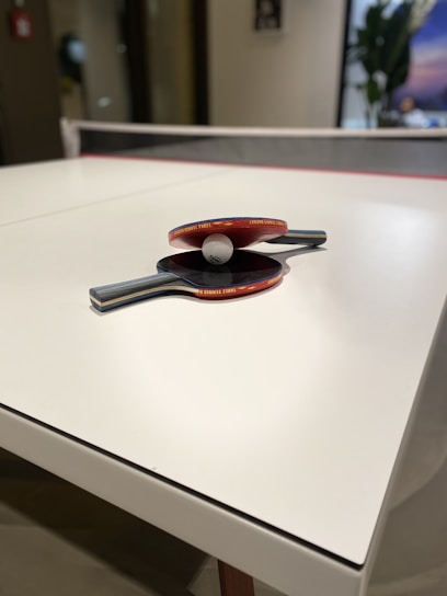 A white table tennis table is seen with two rackets and a ball placed on its surface. The rackets have red rubber surfaces and wooden handles. The ball is small and white, positioned between the two rackets. The setting appears to be indoors with a blurred background that includes a plant and a poster.