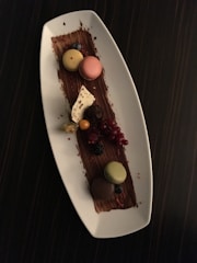 A white rectangular plate displays an elegant dessert arrangement. Three macarons, in shades of yellow, pink, and green, are placed atop a chocolate smear. There are also small fruits like blueberries, redcurrants, and a blackberry. A decorative piece of white chocolate and a chocolate truffle add to the sophisticated presentation, all set against a dark tabletop background.