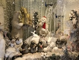A festive display featuring a variety of animal and angel-like figures. A large polar bear holds gold gift boxes, while another wears a Santa hat. In the foreground, small animals like rabbits and what appear to be beavers are arranged among snow-like decorations. Fairy-like figures adorned with wings and winter attire are positioned on the sides. There are decorative trees, fairy lights, and branches covered with artificial snow, creating a whimsical winter scene.