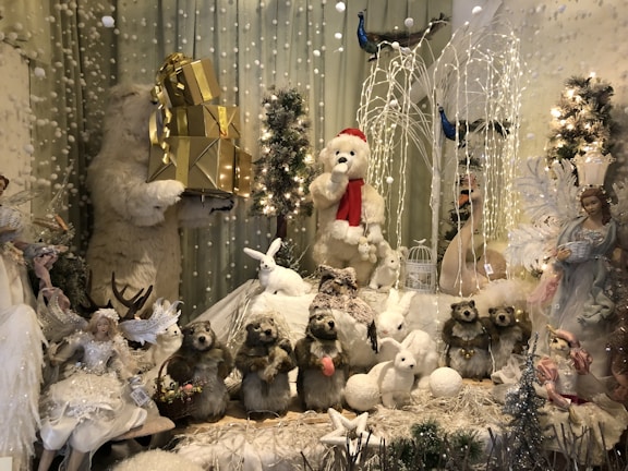 A festive display featuring a variety of animal and angel-like figures. A large polar bear holds gold gift boxes, while another wears a Santa hat. In the foreground, small animals like rabbits and what appear to be beavers are arranged among snow-like decorations. Fairy-like figures adorned with wings and winter attire are positioned on the sides. There are decorative trees, fairy lights, and branches covered with artificial snow, creating a whimsical winter scene.