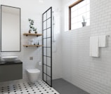 A modern bathroom with white subway tile walls and black and white hexagonal floor tiles. There is a toilet and a floating vanity with a round sink and a large mirror above it. Two wooden shelves hold various decorative items and plants. A shower area with a black-framed glass partition is situated next to a window with a small potted cactus. A towel rack with white towels is mounted on the wall.