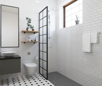 A modern bathroom with white subway tile walls and black and white hexagonal floor tiles. There is a toilet and a floating vanity with a round sink and a large mirror above it. Two wooden shelves hold various decorative items and plants. A shower area with a black-framed glass partition is situated next to a window with a small potted cactus. A towel rack with white towels is mounted on the wall.
