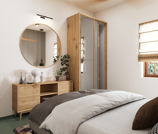 A modern bedroom with minimalist decor, featuring a large round mirror above a wooden dresser with various decorative vases. The bed is neatly made with layered bedding in neutral tones. A potted plant adds a touch of greenery beside the dresser. There are cream-colored Roman shades covering a window, allowing natural light to enter the room.