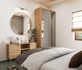A modern bedroom with minimalist decor, featuring a large round mirror above a wooden dresser with various decorative vases. The bed is neatly made with layered bedding in neutral tones. A potted plant adds a touch of greenery beside the dresser. There are cream-colored Roman shades covering a window, allowing natural light to enter the room.