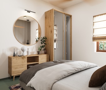 A modern bedroom with minimalist decor, featuring a large round mirror above a wooden dresser with various decorative vases. The bed is neatly made with layered bedding in neutral tones. A potted plant adds a touch of greenery beside the dresser. There are cream-colored Roman shades covering a window, allowing natural light to enter the room.