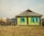 A rustic, single-story house with a corrugated metal roof and bright green walls highlighted by vibrant blue window frames. The home has a weathered, sturdy wooden fence nearby and is surrounded by a dry grassy area. In the background, additional houses and stacks of firewood are visible, giving a sense of rural life.