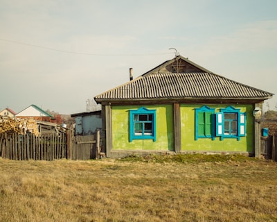 A rustic, single-story house with a corrugated metal roof and bright green walls highlighted by vibrant blue window frames. The home has a weathered, sturdy wooden fence nearby and is surrounded by a dry grassy area. In the background, additional houses and stacks of firewood are visible, giving a sense of rural life.