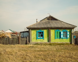 A rustic, single-story house with a corrugated metal roof and bright green walls highlighted by vibrant blue window frames. The home has a weathered, sturdy wooden fence nearby and is surrounded by a dry grassy area. In the background, additional houses and stacks of firewood are visible, giving a sense of rural life.