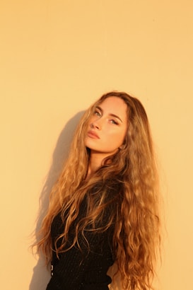 A person with long, curly hair stands against a warm, golden-hued background. The lighting casts a soft glow, highlighting the natural texture and color of the hair. The expression is calm and contemplative.