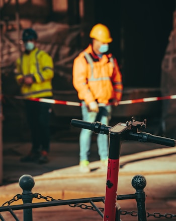 A construction site with two workers in the background wearing protective gear and face masks. One is in a yellow jacket, and the other is in an orange jacket with a hard hat. In the foreground, there is a red electric scooter parked near a chain-linked fence.