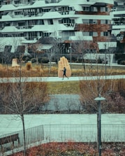 A modern urban scene featuring a runner on a path in a park, with a large, artistic hand sculpture made of brick in the background. The park is surrounded by bushes and bare trees, and a contemporary building with a layered facade stands behind the park.