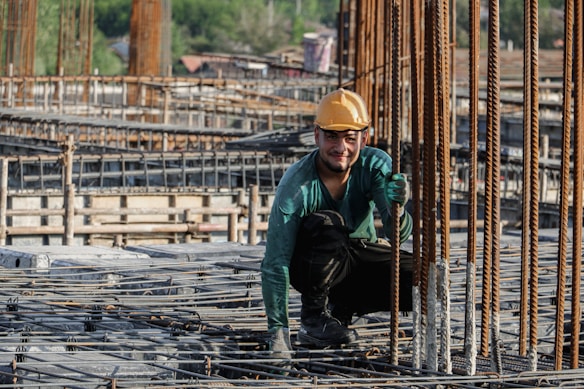 A construction worker wearing a yellow hard hat and green long-sleeve shirt is crouched down on a building site, smiling at the camera. Metal rods and wooden structures surround him, indicating an active construction area.