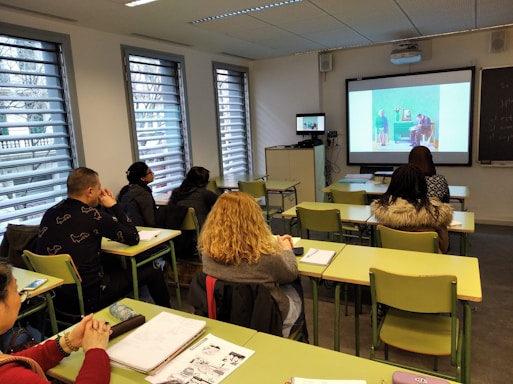 A classroom with several students seated at desks, facing a screen displaying an image. The room has large windows with blinds, and there is a smaller monitor showing the same image as the main screen. The students appear to be focused on the presentation.