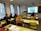 A classroom with several students seated at desks, facing a screen displaying an image. The room has large windows with blinds, and there is a smaller monitor showing the same image as the main screen. The students appear to be focused on the presentation.