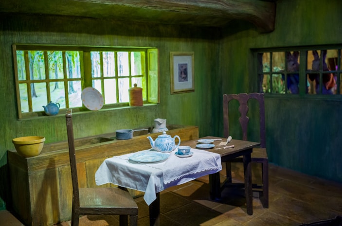 A quaint and rustic dining room features a wooden table with a lace-trimmed white tablecloth. On the table is a blue and white tea set, including a teapot, teacup, and saucers. Two wooden chairs flank the table. A window with a vibrant view of a green forest landscape sits above a sink area, which holds various ceramic kitchen items.