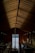 A wooden boathouse interior with a high, triangular ceiling. Wooden beams and panels create an intricate pattern, illuminated by pendant lights. Two boats rest inside the boathouse on calm water, with an exit leading to a lake surrounded by distant trees.
