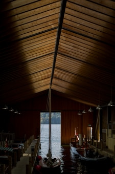 A wooden boathouse interior with a high, triangular ceiling. Wooden beams and panels create an intricate pattern, illuminated by pendant lights. Two boats rest inside the boathouse on calm water, with an exit leading to a lake surrounded by distant trees.