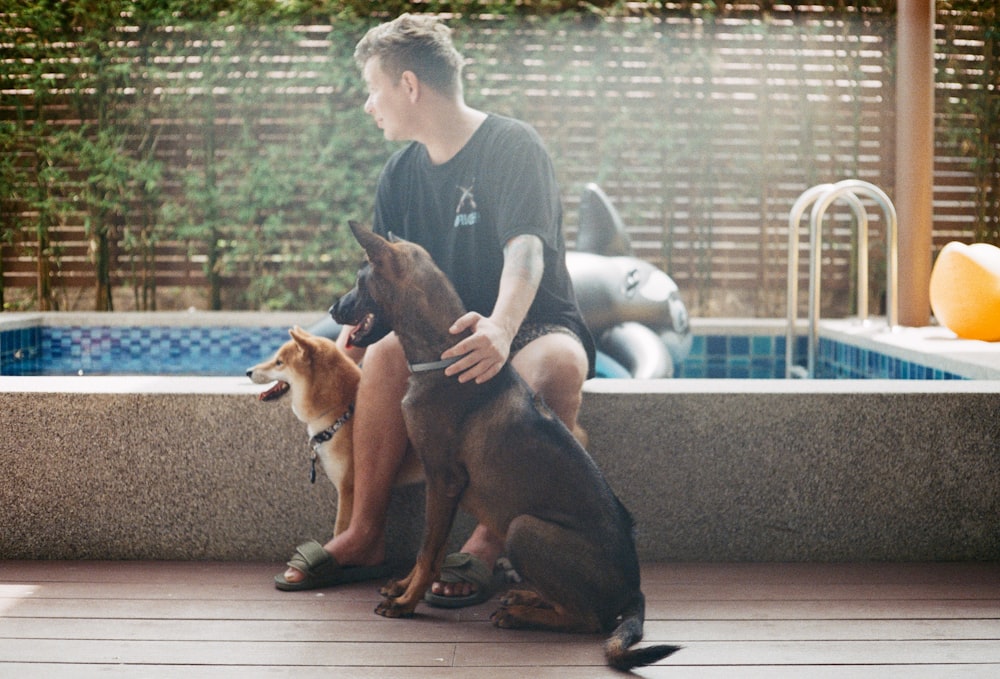 a man sitting next to two dogs in front of a pool