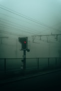 A foggy scene with a railway signal displaying a red light. The signal is mounted on a pole next to train tracks, which are partially obscured by a chain-link fence. Overhead power lines run across the image, contributing to an industrial and mysterious atmosphere.