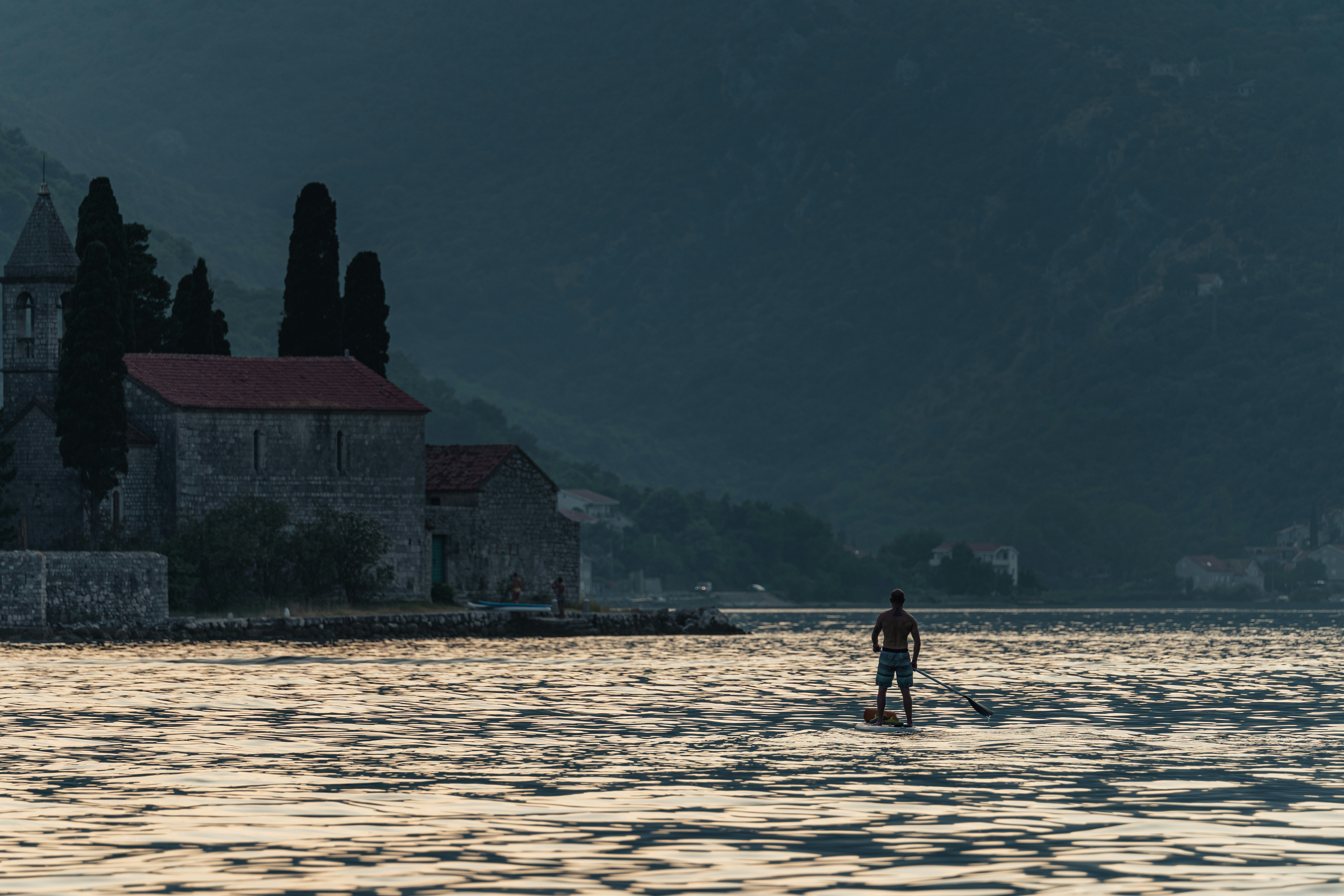 A lone stand-up paddleboarder gliding across the glassy Bay of Kotor at dusk beside the island church of St George