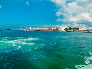 A scenic coastline with vibrant blue waters in the foreground. The shoreline features a mix of buildings, including several with white facades and a prominent lighthouse. The sky is partly cloudy, with patches of blue contrasting against the white clouds.