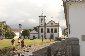 An old colonial-style church stands prominently with white walls and a square bell tower, surrounded by green lawns and stone-paved streets. Several people are walking and socializing in the open space. There is a horse-drawn carriage near the church, and other traditional buildings are visible along the street. The setting features a late afternoon light under a cloudy sky.