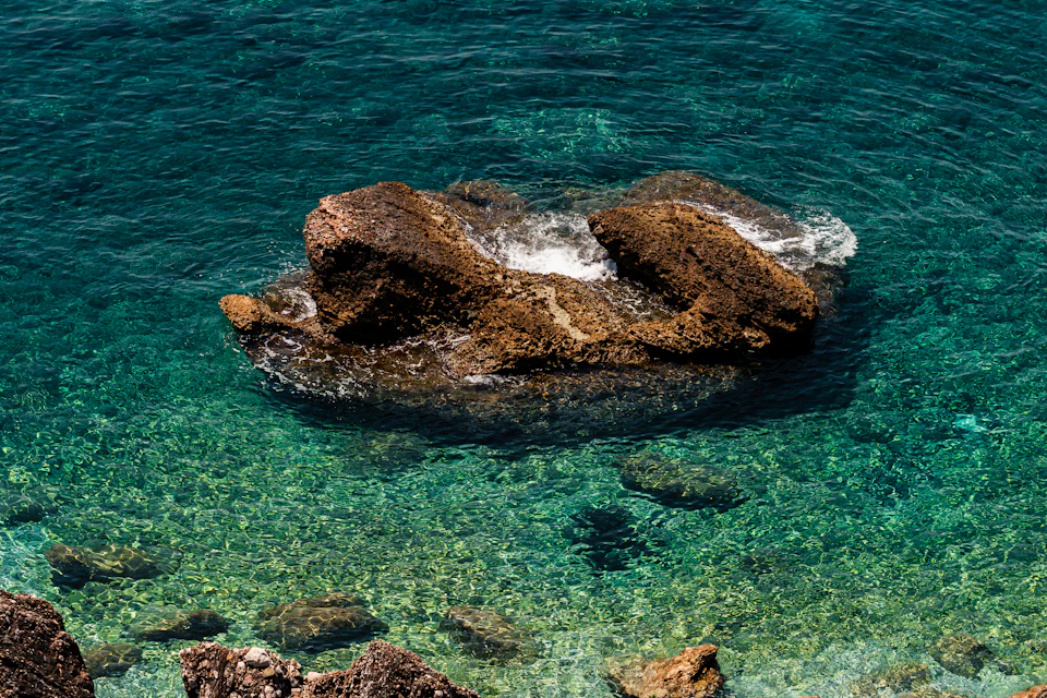 The image depicts a serene underwater scene with a large rock formation in the center, surrounded by clear blue water and small rocks scattered around it. The rock formation appears to be a mix of brown and gray hues, adding depth to the scene. The perspective is from above, providing an expansive view of the rocky island amidst the vast expanse of the ocean.