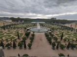 An expansive, meticulously maintained garden features symmetrical designs with geometric and spiral patterns. A large circular fountain sits centrally, surrounded by greenery and carefully trimmed trees. The garden extends into the distance, bordered by tall hedges and stone structures. Overcast skies add a moody ambiance.