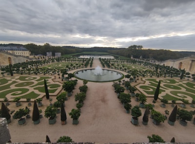 An expansive, meticulously maintained garden features symmetrical designs with geometric and spiral patterns. A large circular fountain sits centrally, surrounded by greenery and carefully trimmed trees. The garden extends into the distance, bordered by tall hedges and stone structures. Overcast skies add a moody ambiance.