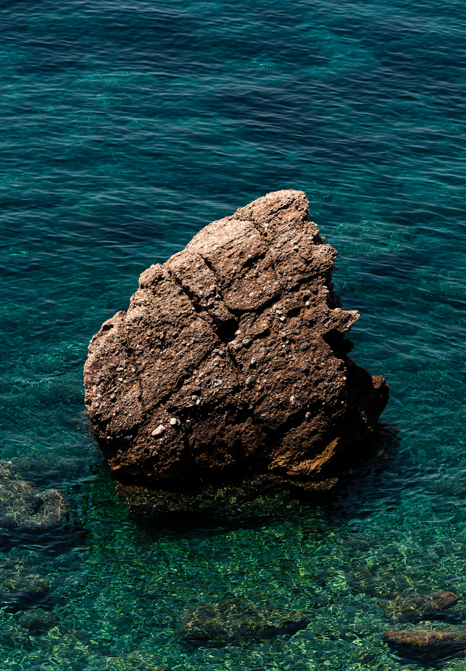 The image depicts a serene scene at sea, featuring a large rock formation with a smooth surface and a small hole on its side. The rock is situated in the center of the frame, surrounded by clear blue water that reflects the light from above. In the background, there are several smaller rocks scattered around the main rock formation, adding depth to the image.