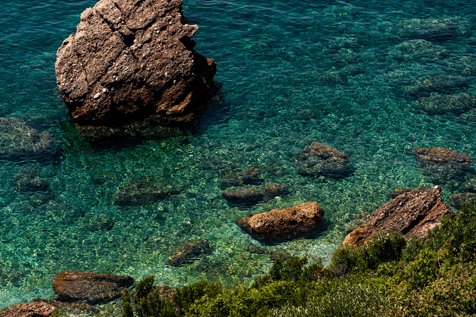The image depicts a serene coastal scene with a large rock formation on the left side, partially submerged in clear blue water and surrounded by small rocks. The deep blue water is dotted with several smaller rocks scattered throughout its surface. On the right side, there are patches of green vegetation, adding a touch of life to the otherwise rocky landscape.