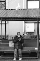 An elderly person sits on a bench under a metal roof while looking at a mobile phone. A large bag is placed next to them on the bench. A seagull stands on the roof above, and a building wall with a utility panel is visible in the background.