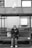 An elderly person sits on a bench under a metal roof while looking at a mobile phone. A large bag is placed next to them on the bench. A seagull stands on the roof above, and a building wall with a utility panel is visible in the background.