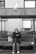 An elderly person sits on a bench under a metal roof while looking at a mobile phone. A large bag is placed next to them on the bench. A seagull stands on the roof above, and a building wall with a utility panel is visible in the background.