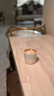 A cup of coffee with latte art sits on a smooth, light-colored marble table. The background is softly blurred, showcasing a cozy interior with chairs and a window with curtains.