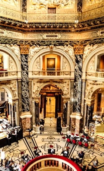 A grand, ornately decorated interior featuring intricate architectural details, statues, and a high ceiling. The floor showcases an elaborate black and white pattern, with dining tables and red chairs arranged for visitors. People are seated, enjoying their meals and beverages, while a bustling counter area provides service.