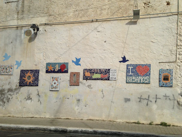 A sunlit wall adorned with a large mosaic depicting traditional Mani landscapes.