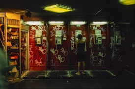 A person stands in front of a row of vintage payphones, each mounted on a wall covered with graffiti. The setting is dimly lit with fluorescent lighting, creating a moody atmosphere. The individual appears to be using one of the phones, and is dressed in a casual sleeveless top and shorts.