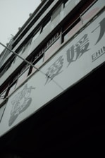 A modern building facade with large glass windows and signage containing Japanese characters. The structure appears to be multi-storied with visible structural lines and a muted color palette.