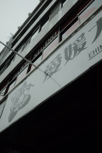 A modern building facade with large glass windows and signage containing Japanese characters. The structure appears to be multi-storied with visible structural lines and a muted color palette.