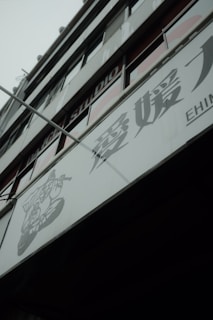 A modern building facade with large glass windows and signage containing Japanese characters. The structure appears to be multi-storied with visible structural lines and a muted color palette.