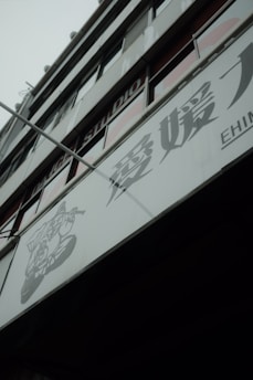 A modern building facade with large glass windows and signage containing Japanese characters. The structure appears to be multi-storied with visible structural lines and a muted color palette.