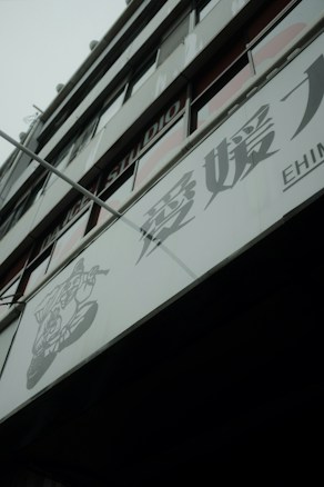 A modern building facade with large glass windows and signage containing Japanese characters. The structure appears to be multi-storied with visible structural lines and a muted color palette.