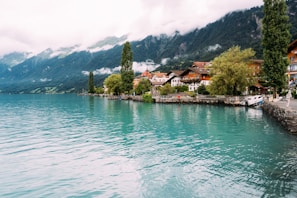 A picturesque lakeside town with charming buildings nestled against a backdrop of lush green mountains. The calm turquoise water of the lake reflects the surrounding scenery, while a few tall trees line the shoreline. The sky is partially cloudy, adding to the serene and tranquil atmosphere.