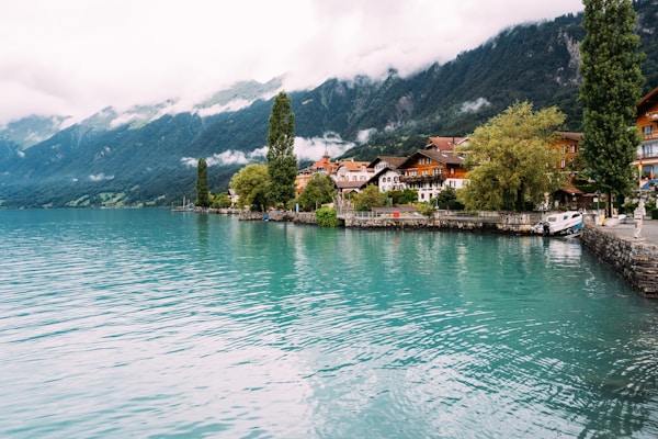 A picturesque lakeside town with charming buildings nestled against a backdrop of lush green mountains. The calm turquoise water of the lake reflects the surrounding scenery, while a few tall trees line the shoreline. The sky is partially cloudy, adding to the serene and tranquil atmosphere.