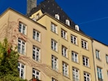 A multi-story building with a light beige facade features numerous windows organized in a repeated pattern. Ivy drapes across part of the building, adding a touch of greenery and red leaves. The roof is dark gray and steeply angled, topped with a chimney. A clear blue sky serves as the backdrop.