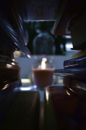 A lit candle is positioned at the center, surrounded by a dark, cozy atmosphere. Soft, warm light emanates from the candle, creating subtle reflections on nearby surfaces. The background features blurred details, enhancing the focus on the candle.
