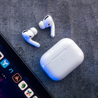 Two wireless earbuds and a charging case placed on a textured surface. A portion of a tablet screen displaying several app icons is visible in the bottom left corner.