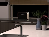 A modern kitchen countertop featuring a metal sink, a silver faucet, and a sleek black toaster. A vase with purple flowers and a green plant in a white pot are also placed on the counter. Blinds cover a window in the background.