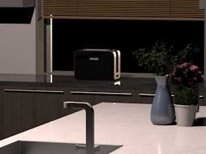A modern kitchen countertop featuring a metal sink, a silver faucet, and a sleek black toaster. A vase with purple flowers and a green plant in a white pot are also placed on the counter. Blinds cover a window in the background.